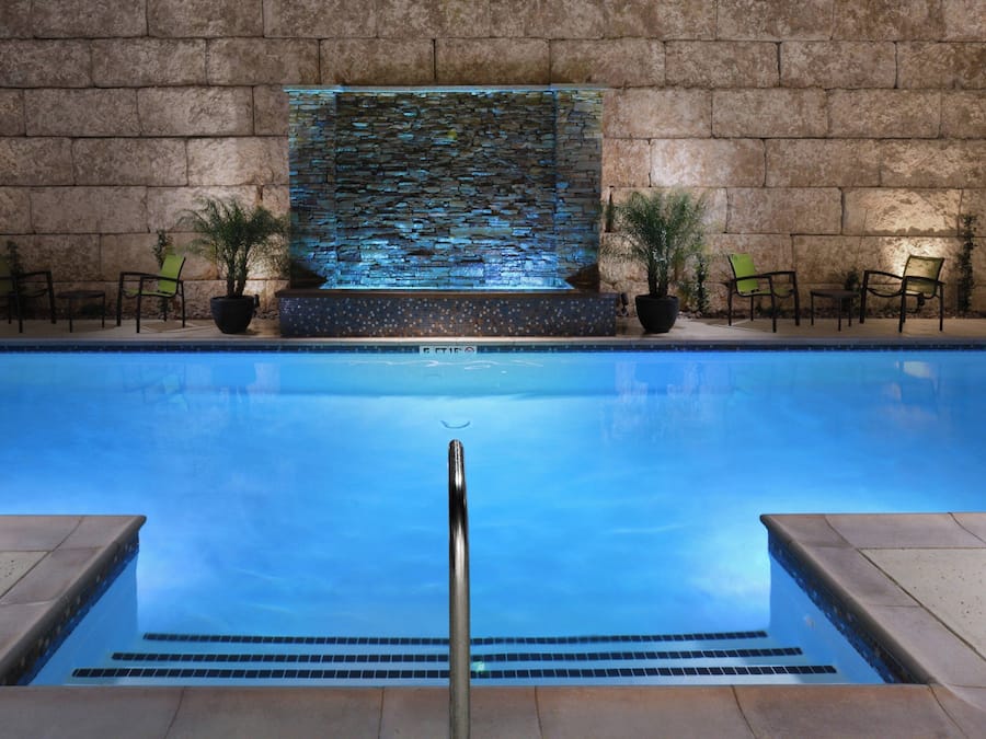 A swimming pool with a stone wall backdrop, a water feature, and potted plants.