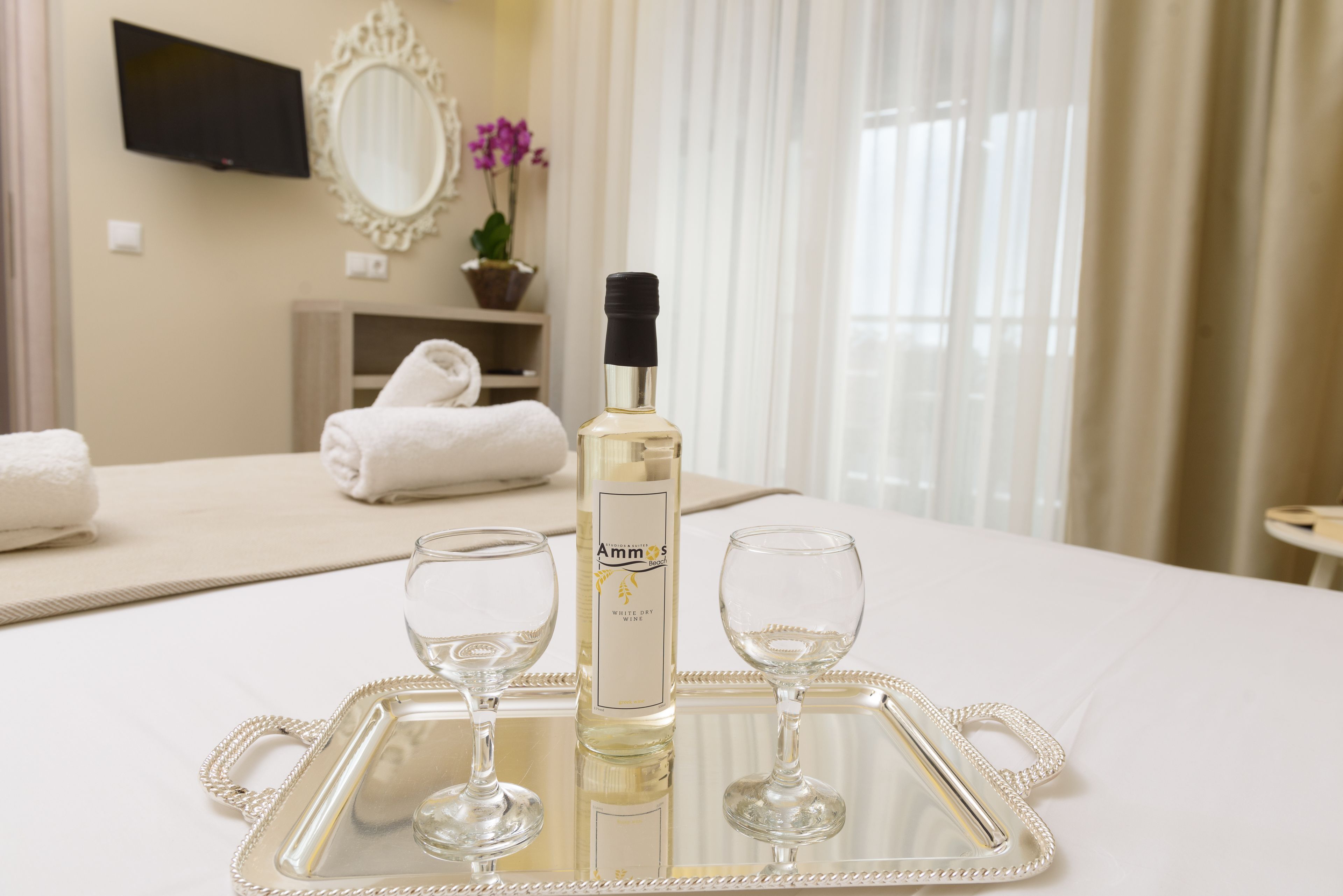 A hotel room with a bed, a tray with two wine glasses, a bottle of wine, a TV, a mirror, and a potted plant.