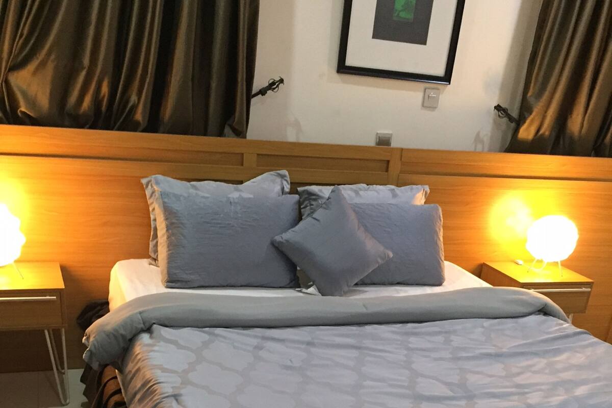 Cama bem arrumada com colcha estampada e travesseiros, com duas luminárias de cabeceira sobre mesas de cabeceira de madeira.