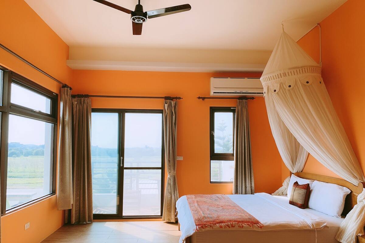 A bedroom with a bed, an air conditioning unit, a ceiling fan, and a view through the window.