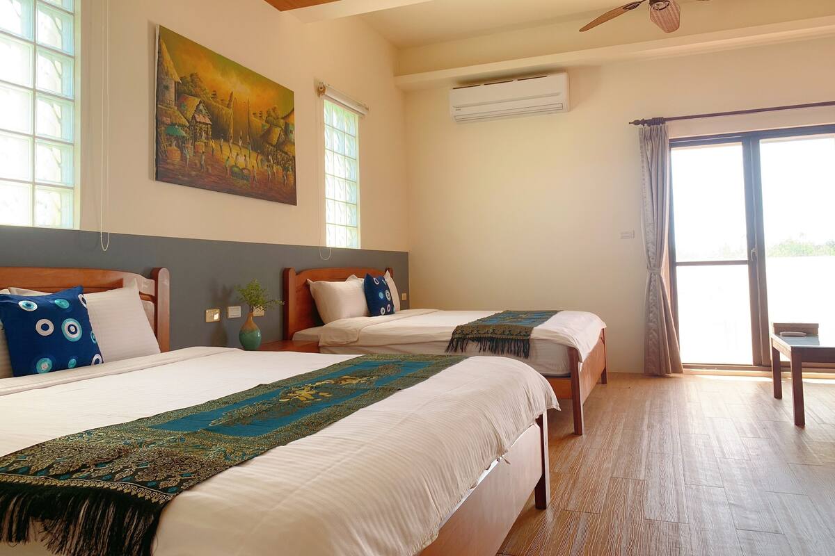 A hotel room with two beds, a ceiling fan, an air conditioning unit, and a large window with curtains.