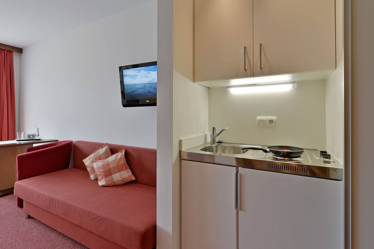 Une chambre d’hôtel compacte comprenant un coin cuisine, un canapé, une petite table et une fenêtre avec des rideaux.
