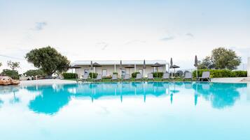 Une piscine avec des chaises longues et des parasols, un bâtiment à la façade blanche, et de la verdure en arrière-plan.