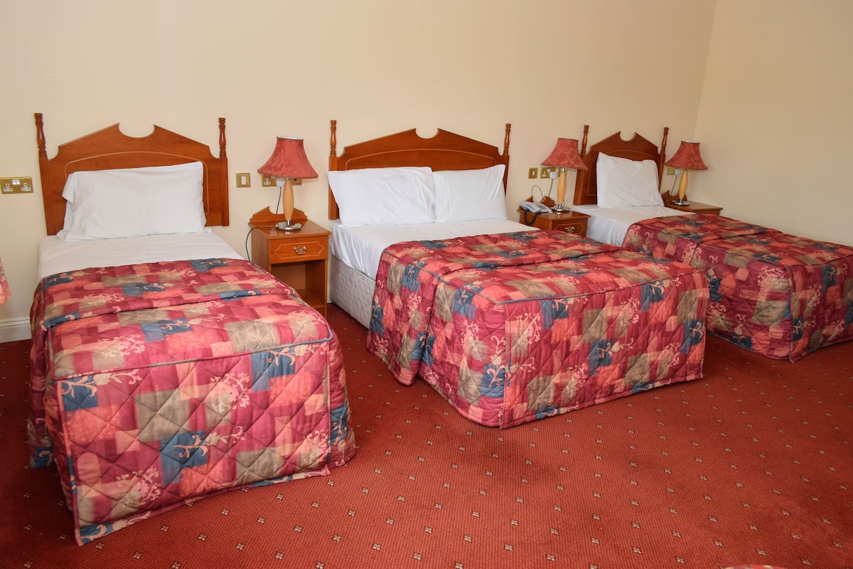 A hotel room with three beds, each with a wooden headboard and floral bedding. There are bedside tables with lamps and a telephone.