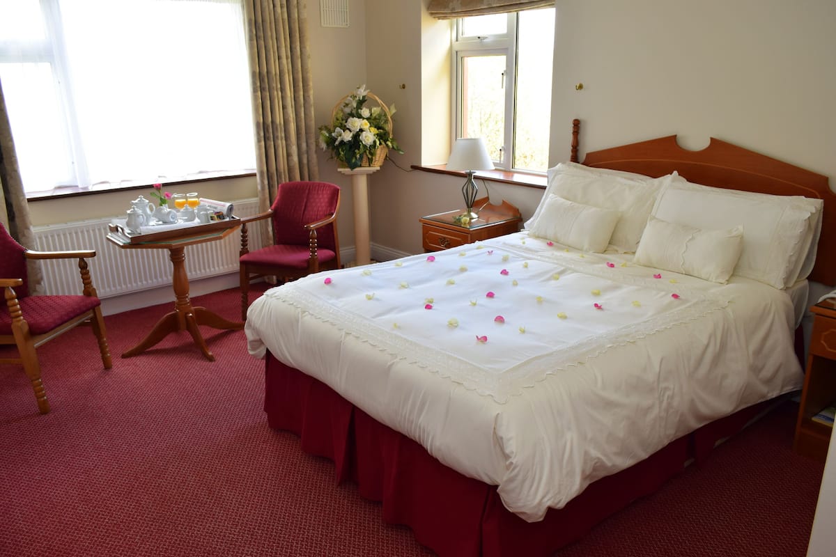 A hotel room with a large bed, a desk with a chair, a window with curtains, and a nightstand with a lamp.