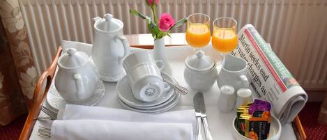 A breakfast tray with white ceramic dishes, a teapot, a sugar bowl, a knife, a fork, a napkin, a vase with a pink rose, two glasses of orange juice, and a newspaper.