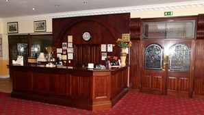 Un comptoir d’accueil avec une horloge, des photos encadrées et un vase rempli de fleurs. Un panneau indique les numéros des chambres, du 20 au 31.