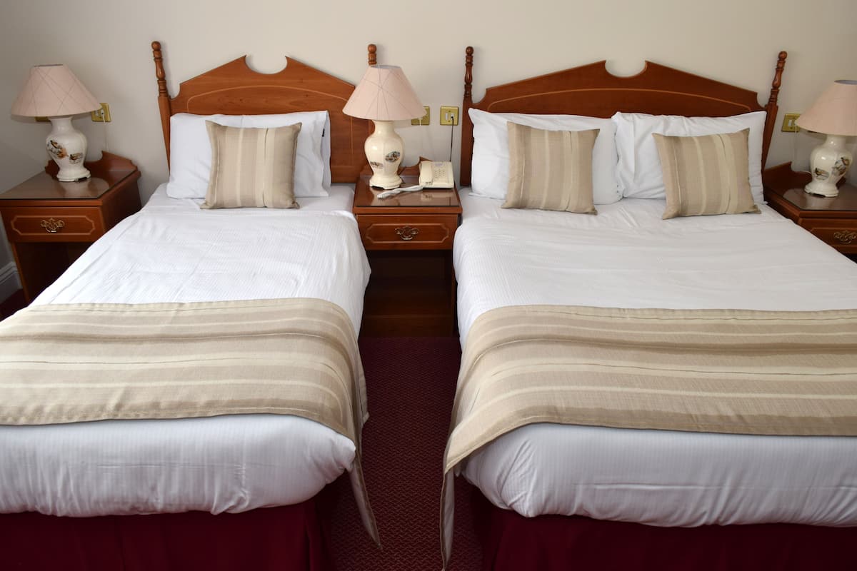 A hotel room with two single beds, each with a wooden headboard, white bedding, and a red carpet. There are bedside tables with lamps and a telephone on each bed.