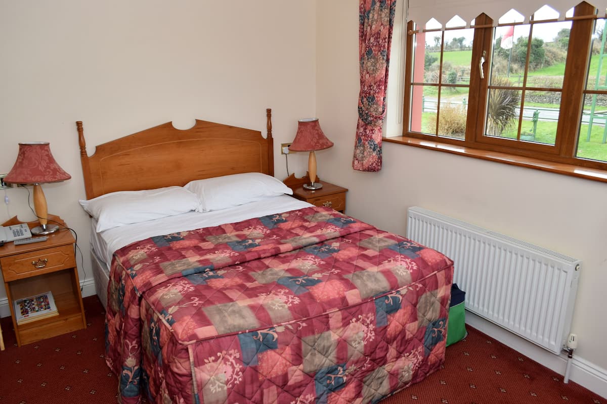 A bedroom with a wooden bed, two bedside tables, a red quilt, a window with a view of greenery, and a radiator.