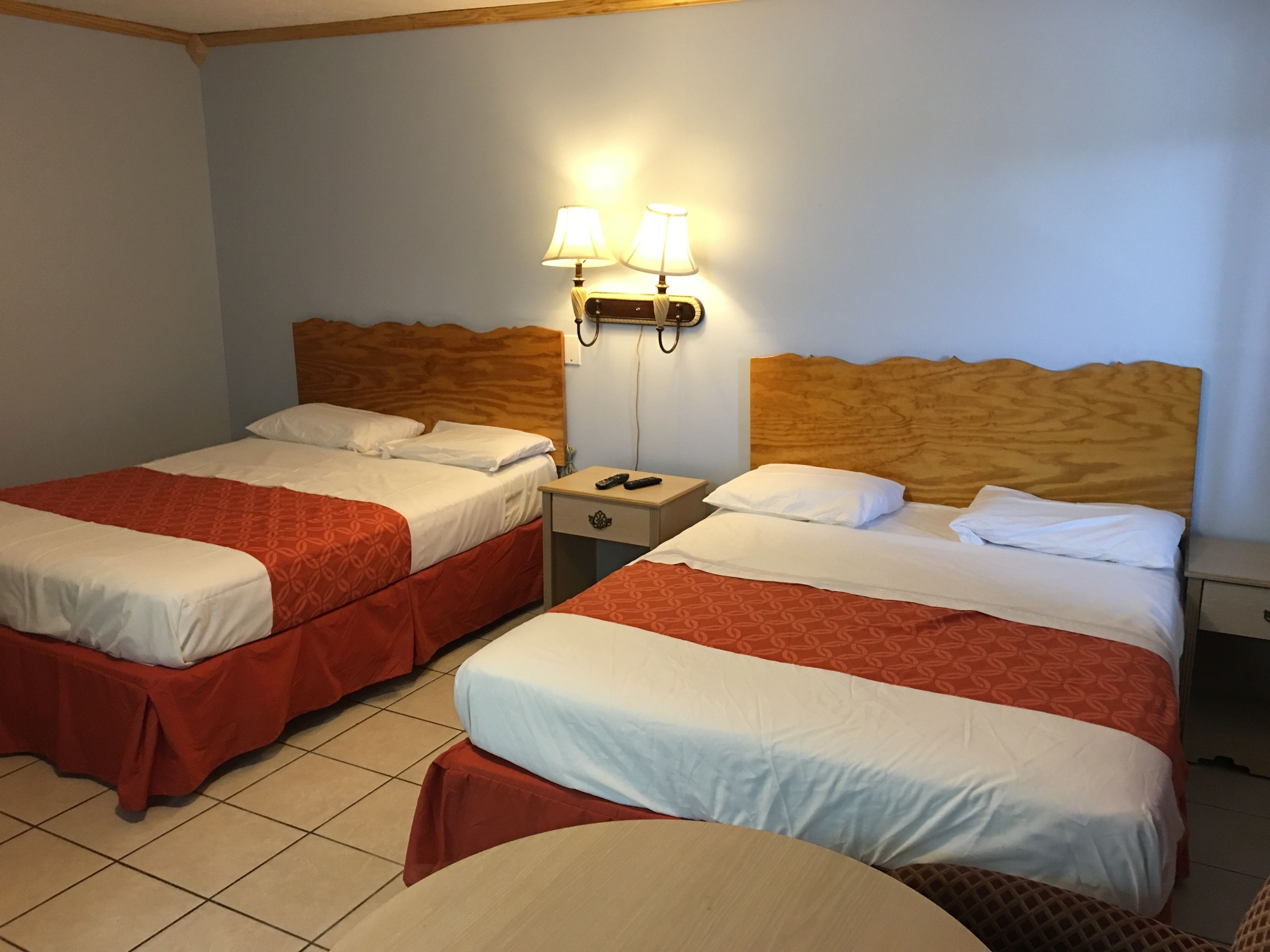 Ein Hotelzimmer mit zwei Betten, jeweils mit Holz-Kopfteilen, weißen Laken und roten Bettdecken. Zwei Nachttische mit Lampen, ein kleiner Tisch und ein Sessel sind ebenfalls vorhanden.