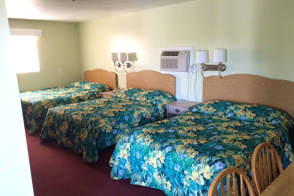 A room with three beds, each with floral bedding, a wall-mounted air conditioner, and bedside lamps.