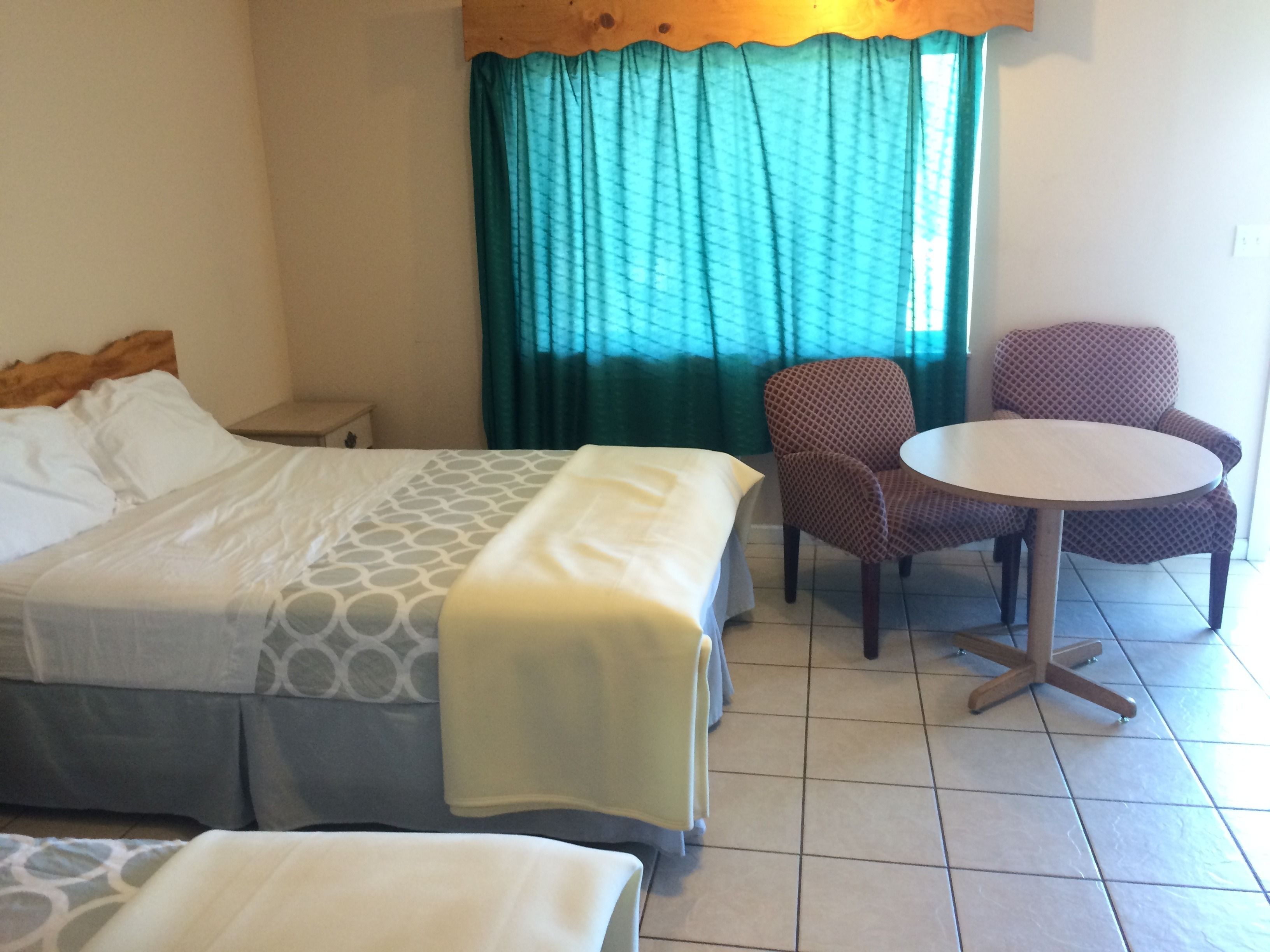 Una habitación de hotel con cama, una mesita, dos sillas y un ventanal con cortinas.