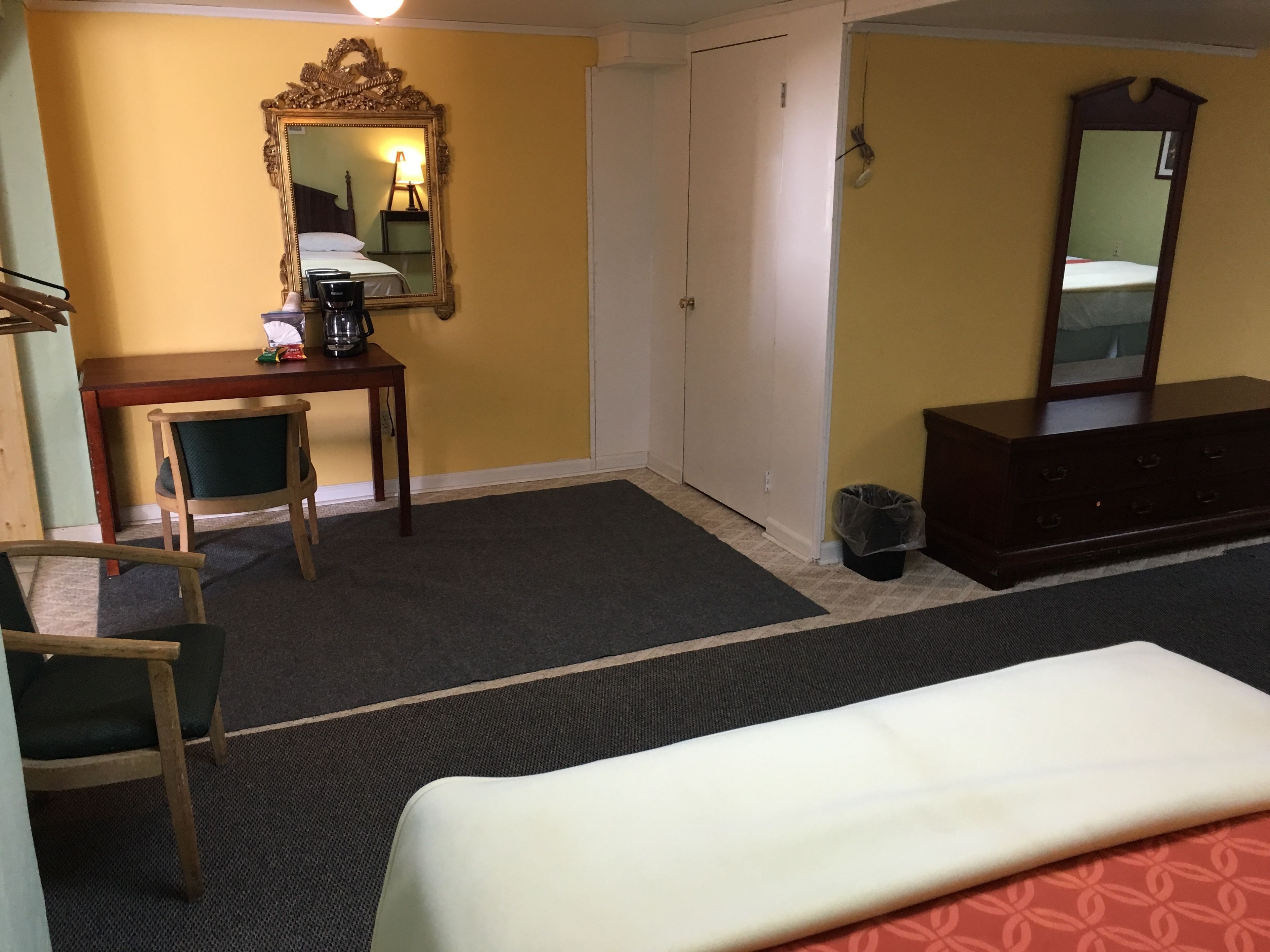 A hotel room with a bed, a desk with a chair, a dresser, a mirror, and a trash can.