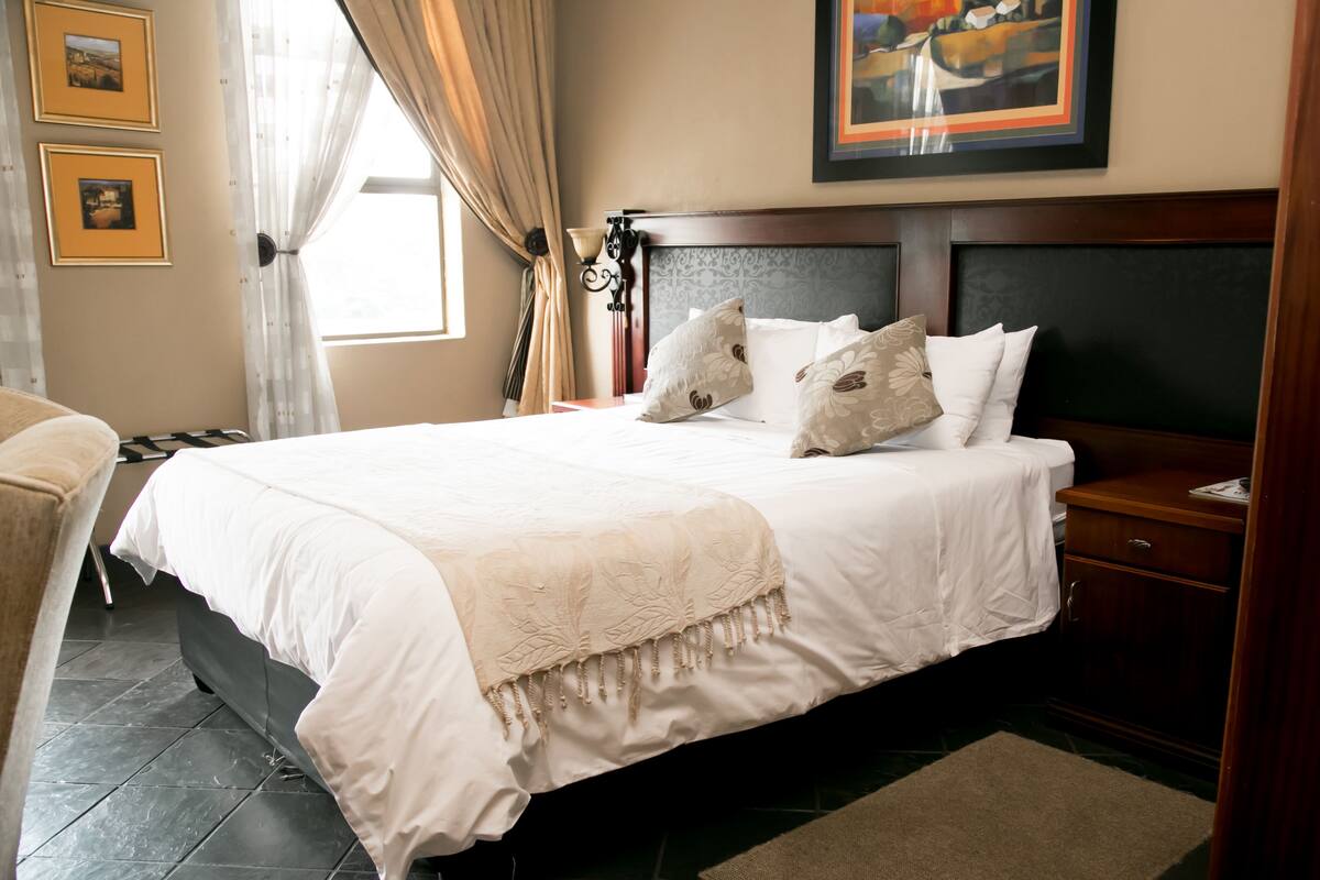 A neatly made bed with a white comforter and pillows, a wooden headboard, a bedside table with a lamp, and a framed picture on the wall.
