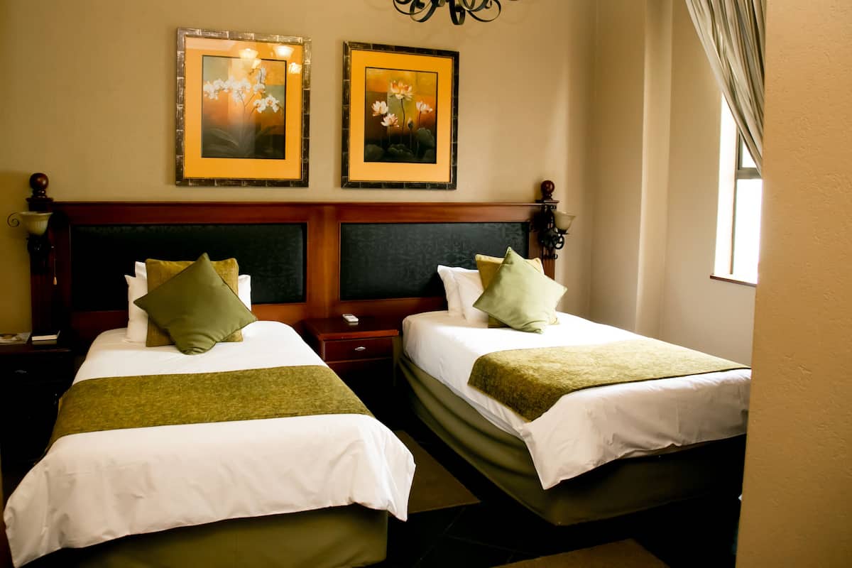 A hotel room with two beds, each with white linens and green throw blankets. There are two framed floral paintings on the wall above the beds.