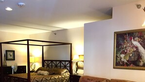 Uma cama com dossel, um ventilador, um sofá marrom, um banquinho de couro e um quadro na parede.