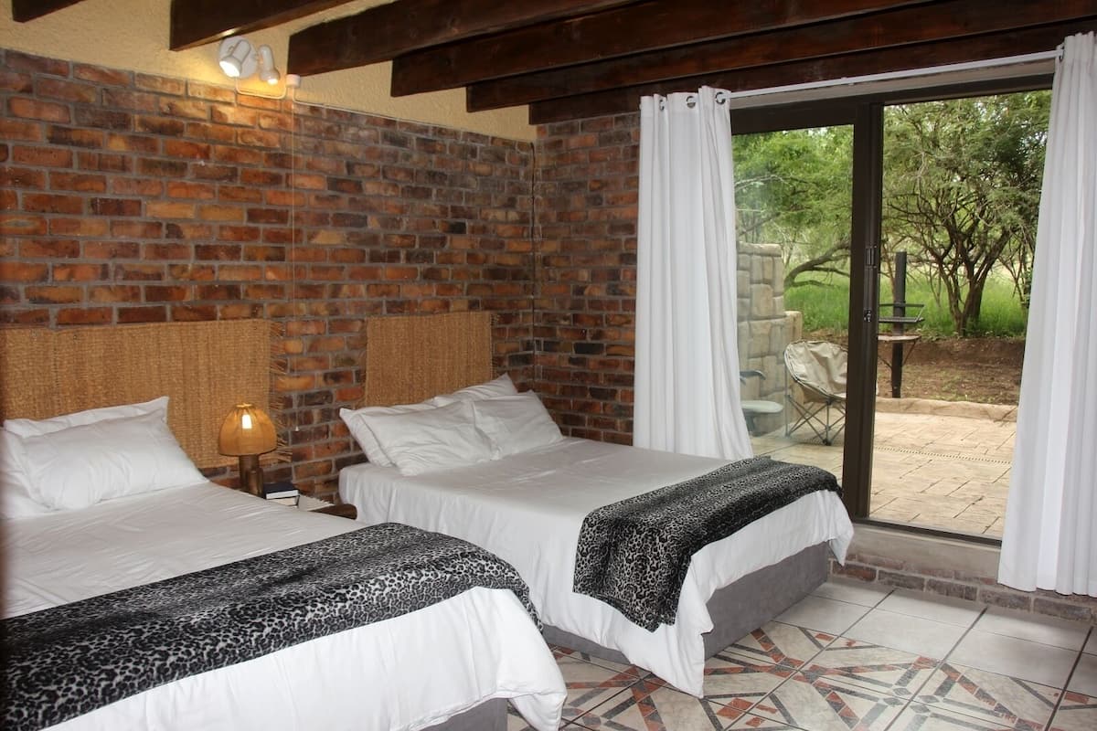 A room with two beds, a brick wall, a woven headboard, a nightstand with a lamp, and a view of an outdoor patio with a table and chairs.