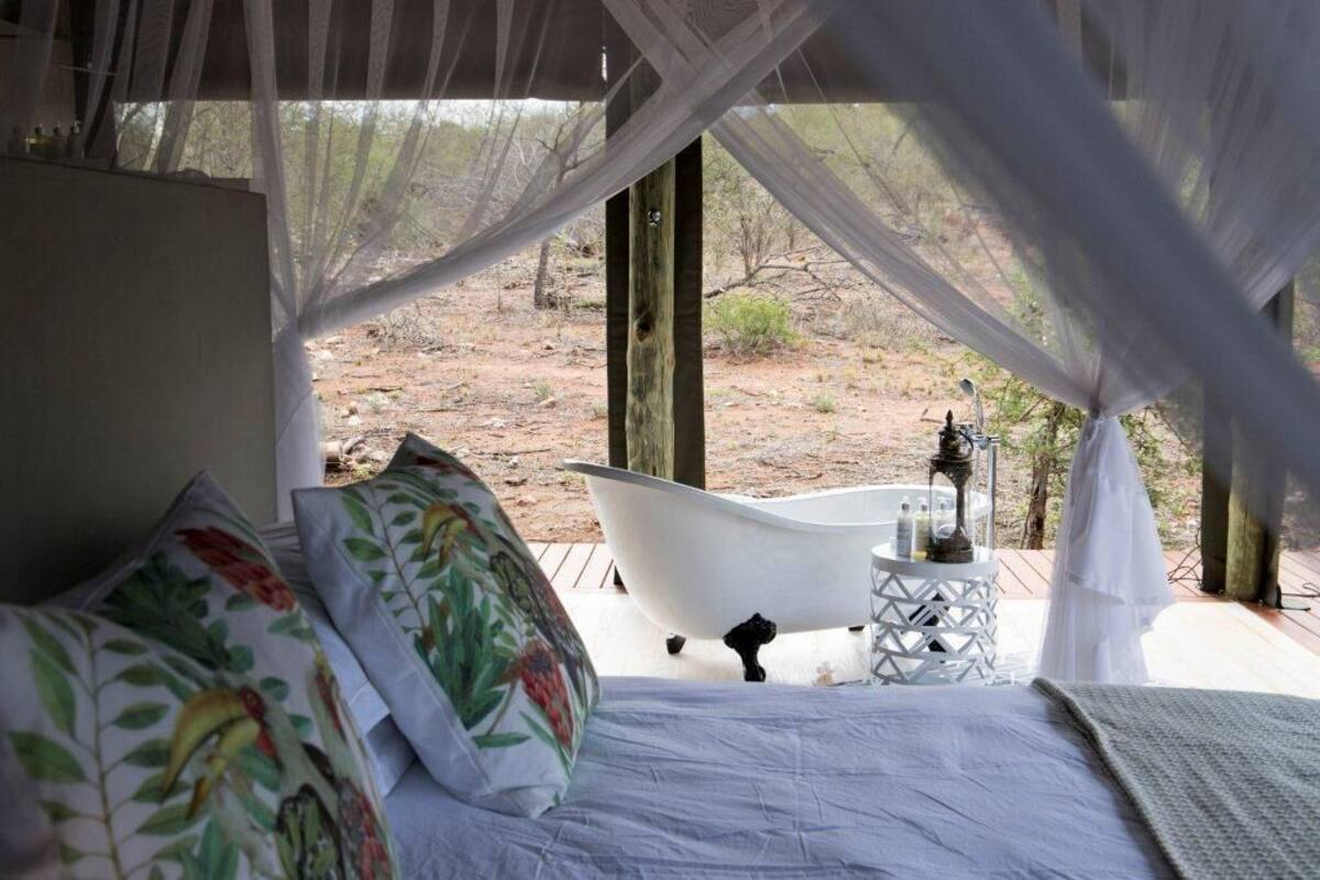 A bedroom with a bed, floral pillows, a white bathtub, and a small table with toiletries.