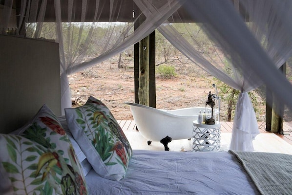 A bedroom with a bed, floral pillows, a white bathtub, and a small table with toiletries.