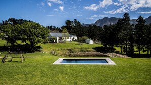 A pool in a grassy area with a house and mountains in the background. - High Season Farm (Hermanus)