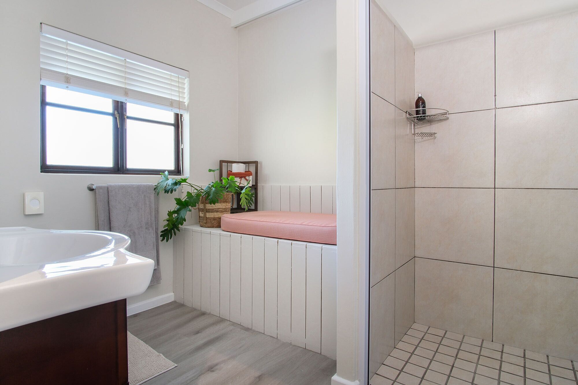 Une salle de bain avec un lavabo blanc, une douche aux carreaux beiges, une fenêtre avec des stores et une plante posée sur le radiateur.