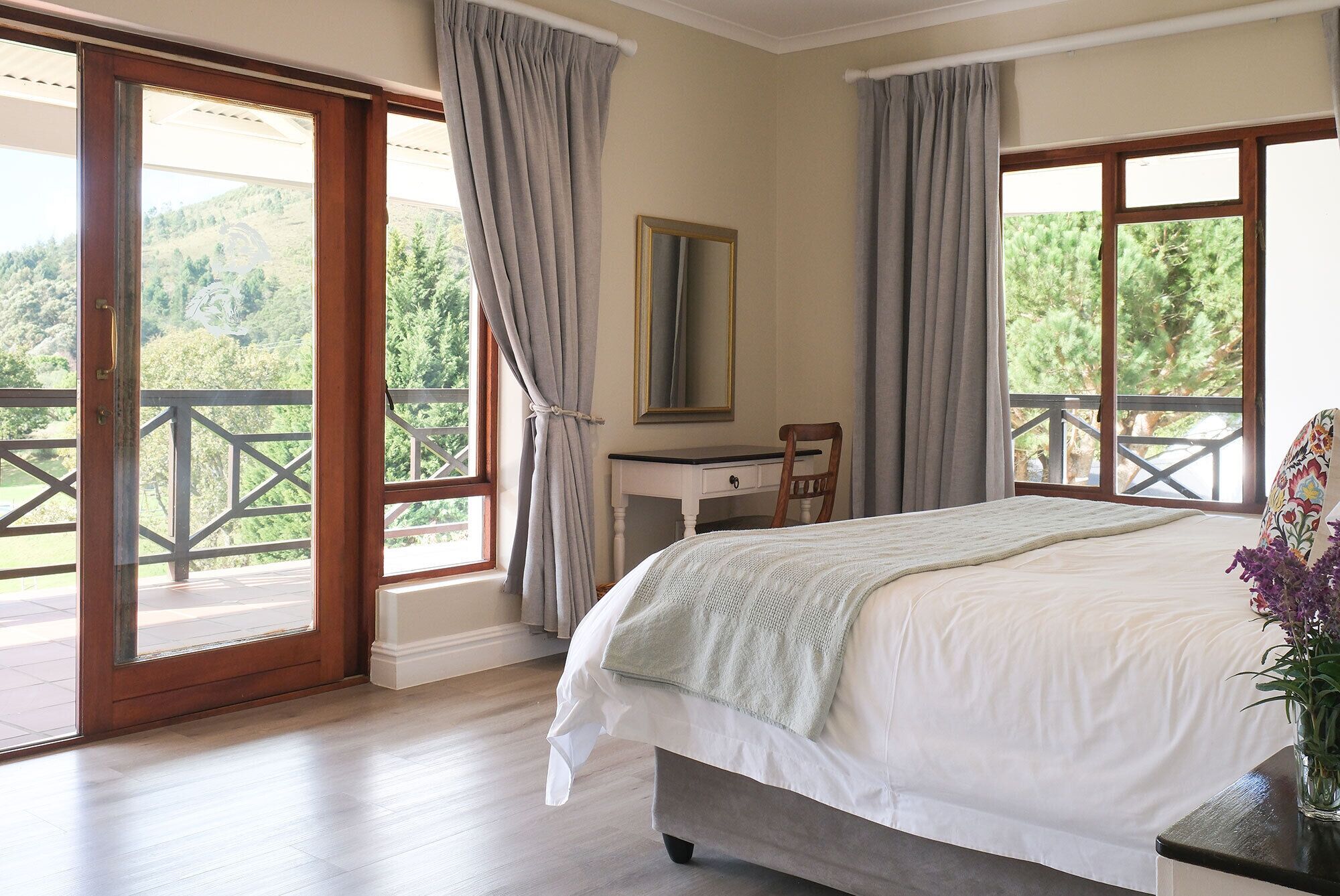 A bedroom with a bed, a desk, and a chair. There are two windows with curtains, and a balcony is visible through one window.