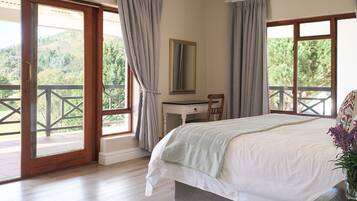 A bedroom with a bed, a desk, and a chair. There are two windows with curtains, and a balcony is visible through one window.