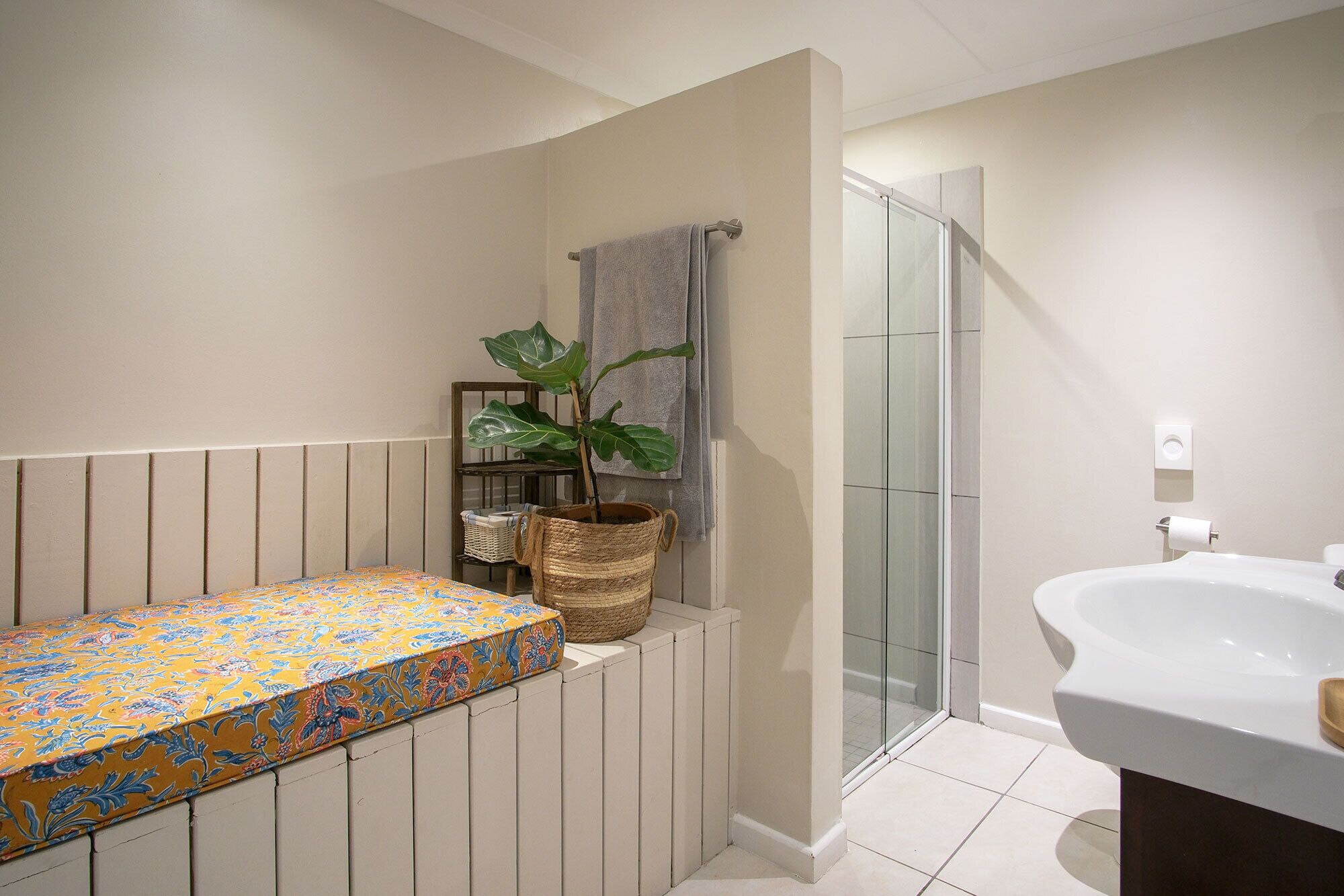 Une salle de bain avec une douche, un lavabo, un banc recouvert d’un tissu à motifs floraux et une plante en pot.