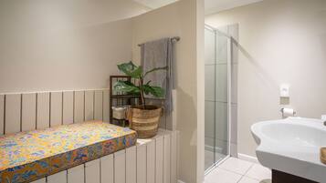 Une salle de bain avec une douche, un lavabo, un banc recouvert d’un tissu à motifs floraux et une plante en pot.