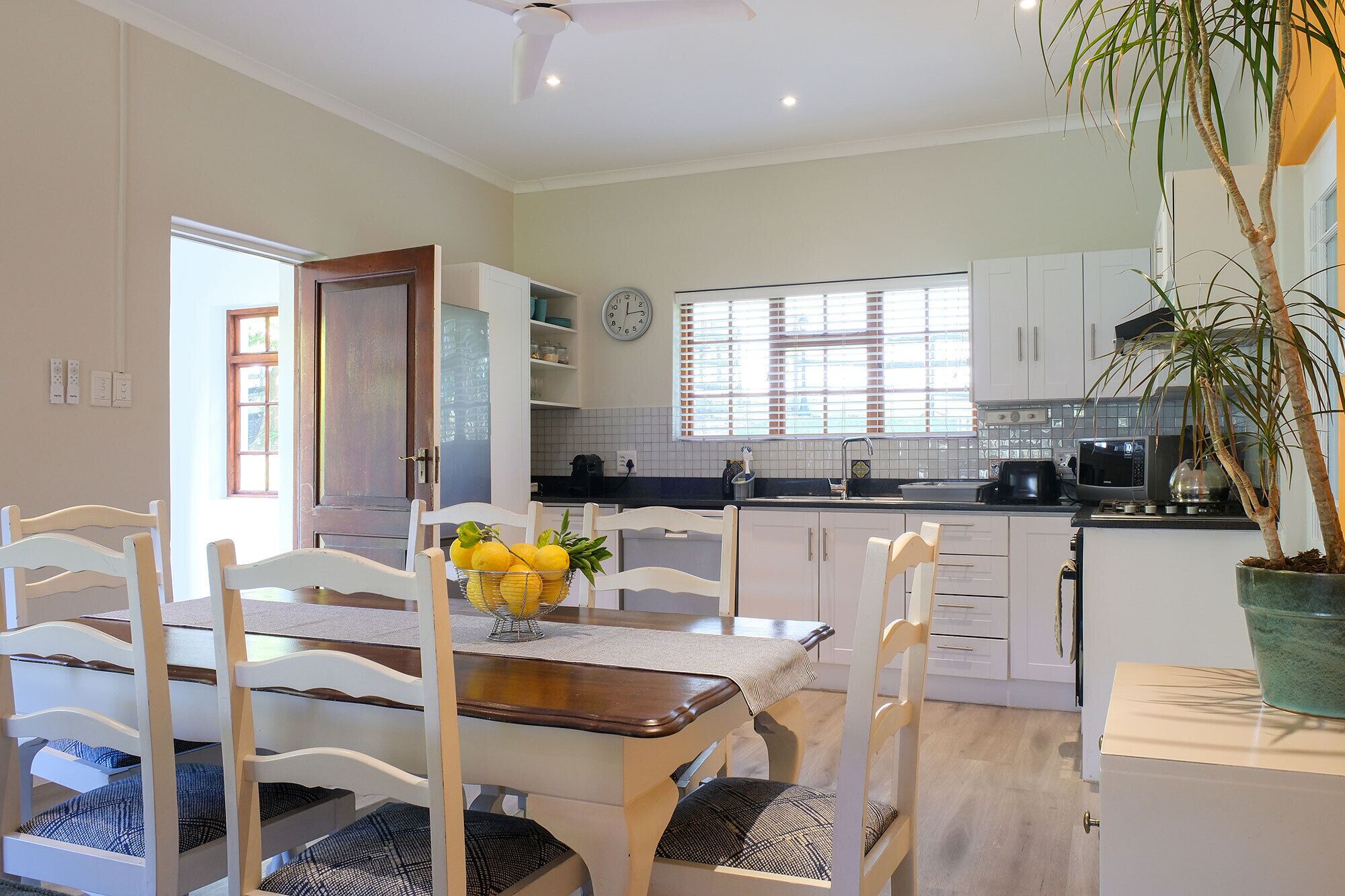 Uma cozinha moderna com armários brancos, uma área de jantar com mesa e cadeiras de madeira, e uma planta em vaso.