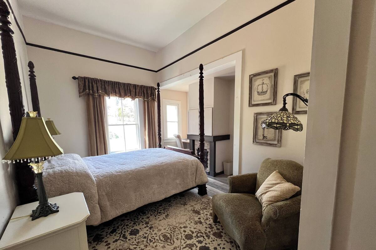 A bedroom with a four-poster bed, a chair, a desk, and a patterned rug.