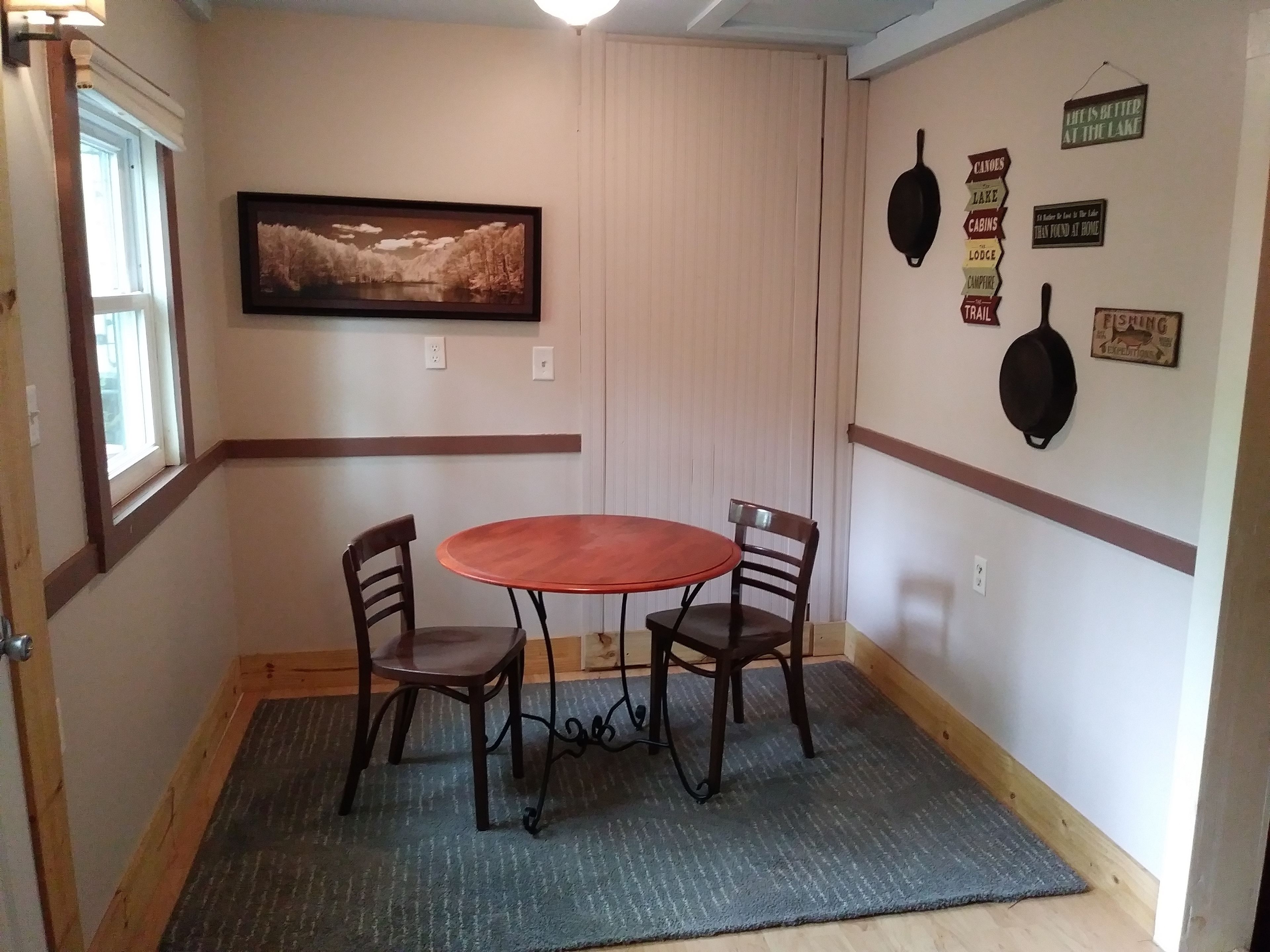 Un comedor con una mesa redonda y dos sillas, un cuadro de paisaje enmarcado en la pared, y varios carteles colocados en la pared relacionados con actividades al aire libre.
