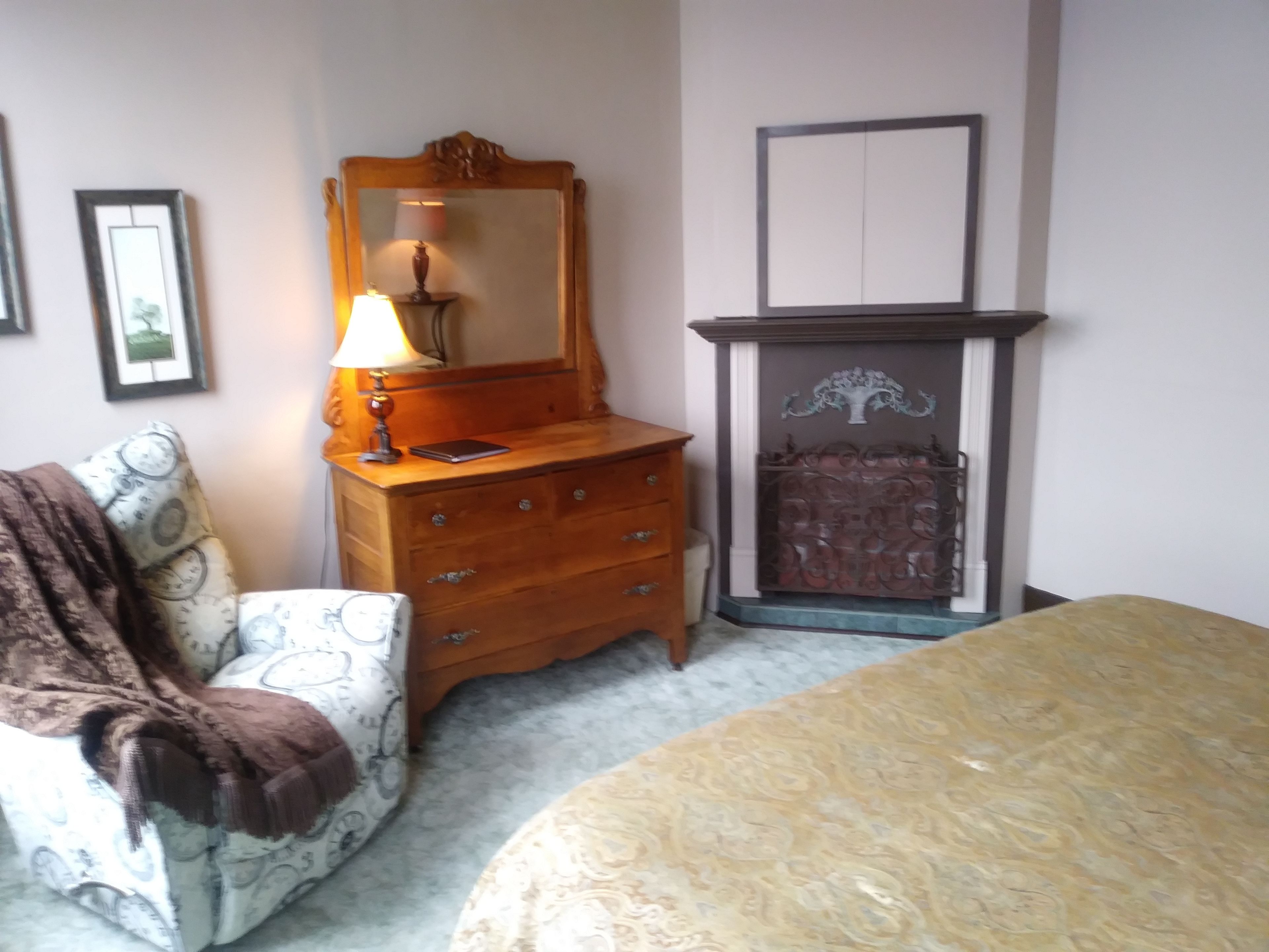 A bedroom with a bed, a dresser, a mirror, a lamp, a fireplace, and a patterned armchair.