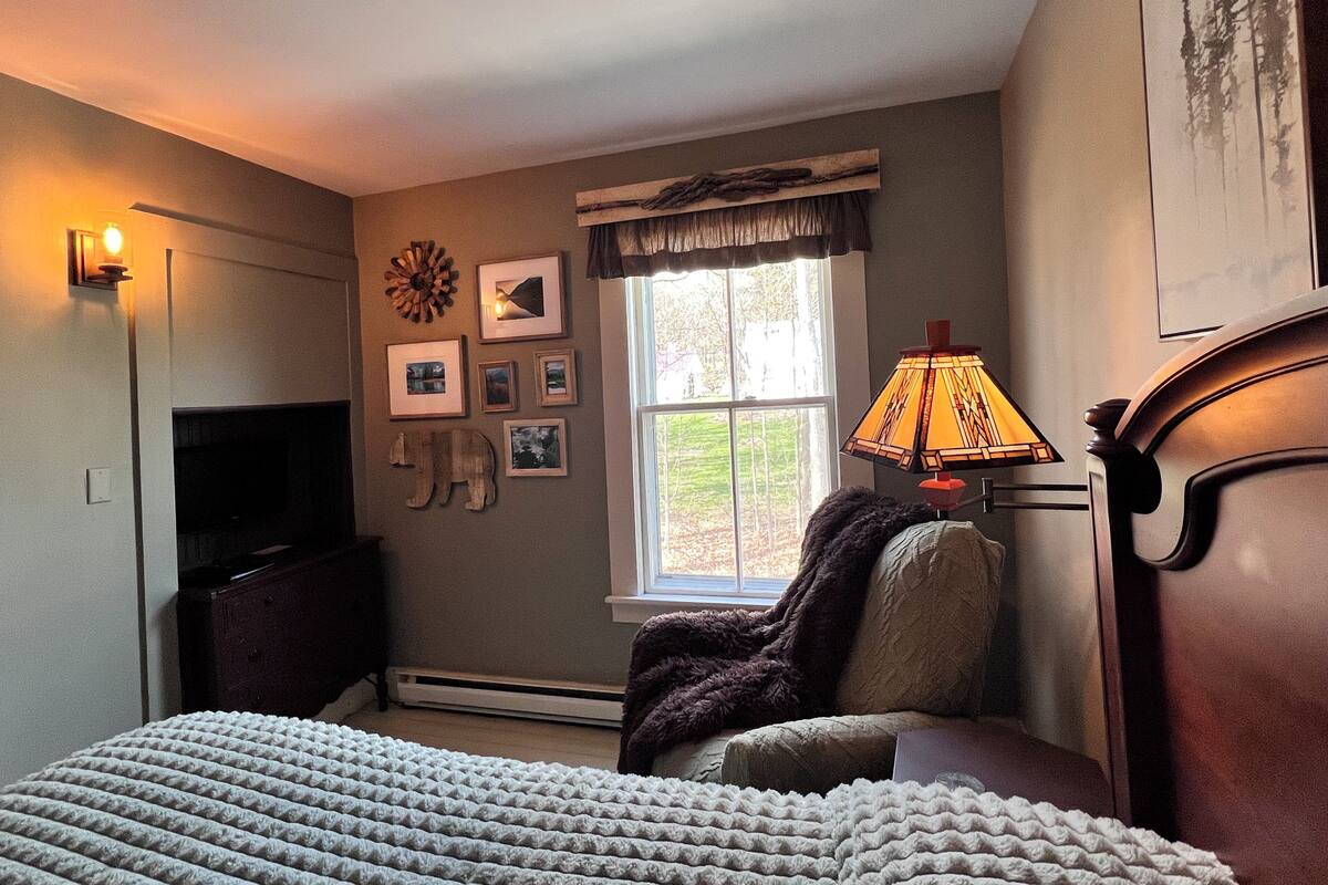A bedroom with a bed, a chair, a lamp, a fireplace, and a window with a view of trees.