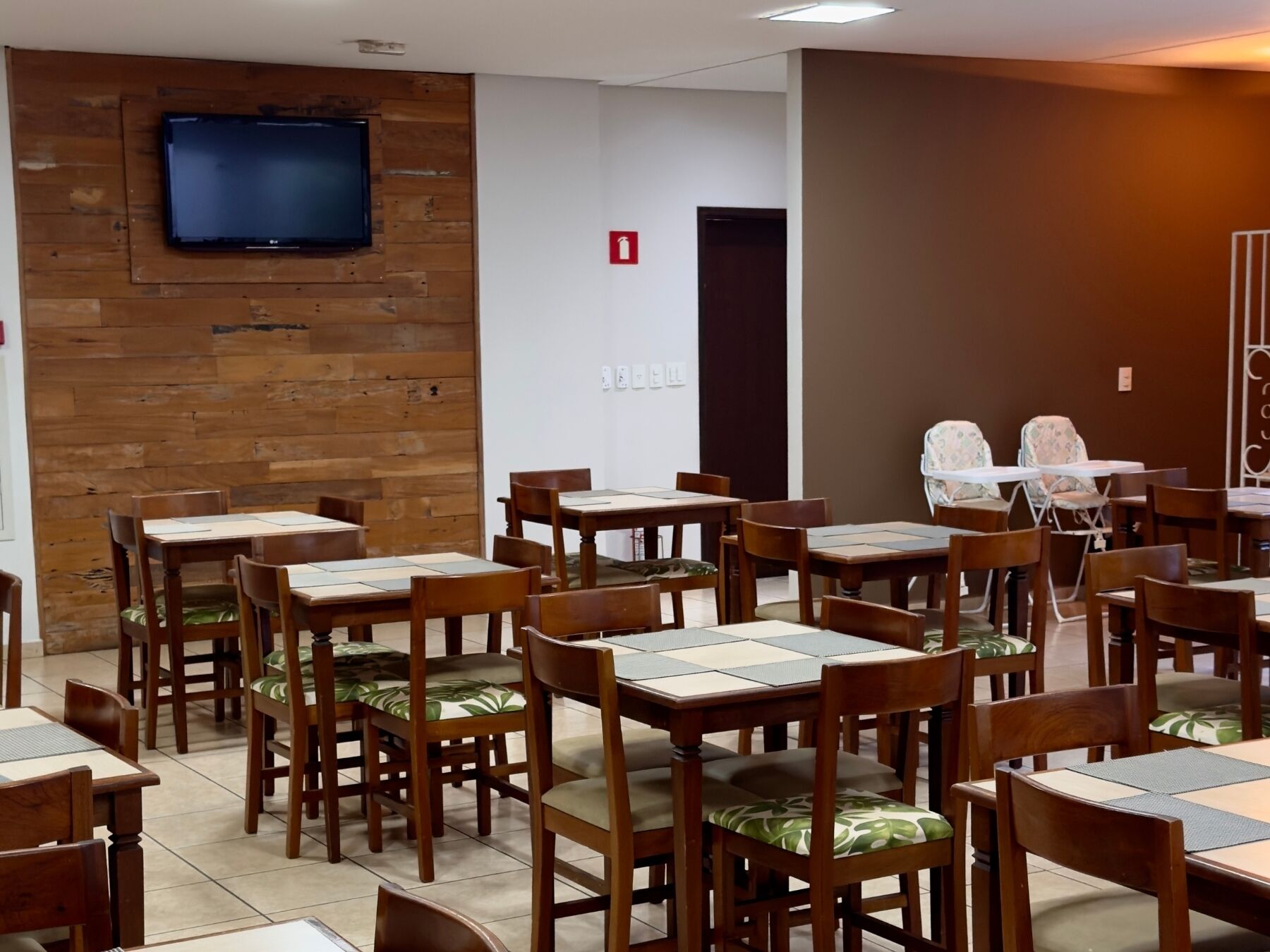 Un comedor con mesas y sillas de madera, un televisor montado en una pared de madera, y un letrero de salida de incendios en la pared.