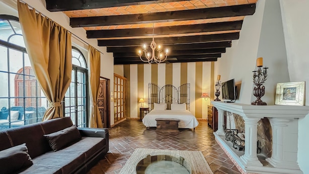 A room with a bed, a sofa, a fireplace, and a large window with curtains. - Casa Liza (San Miguel de Allende)