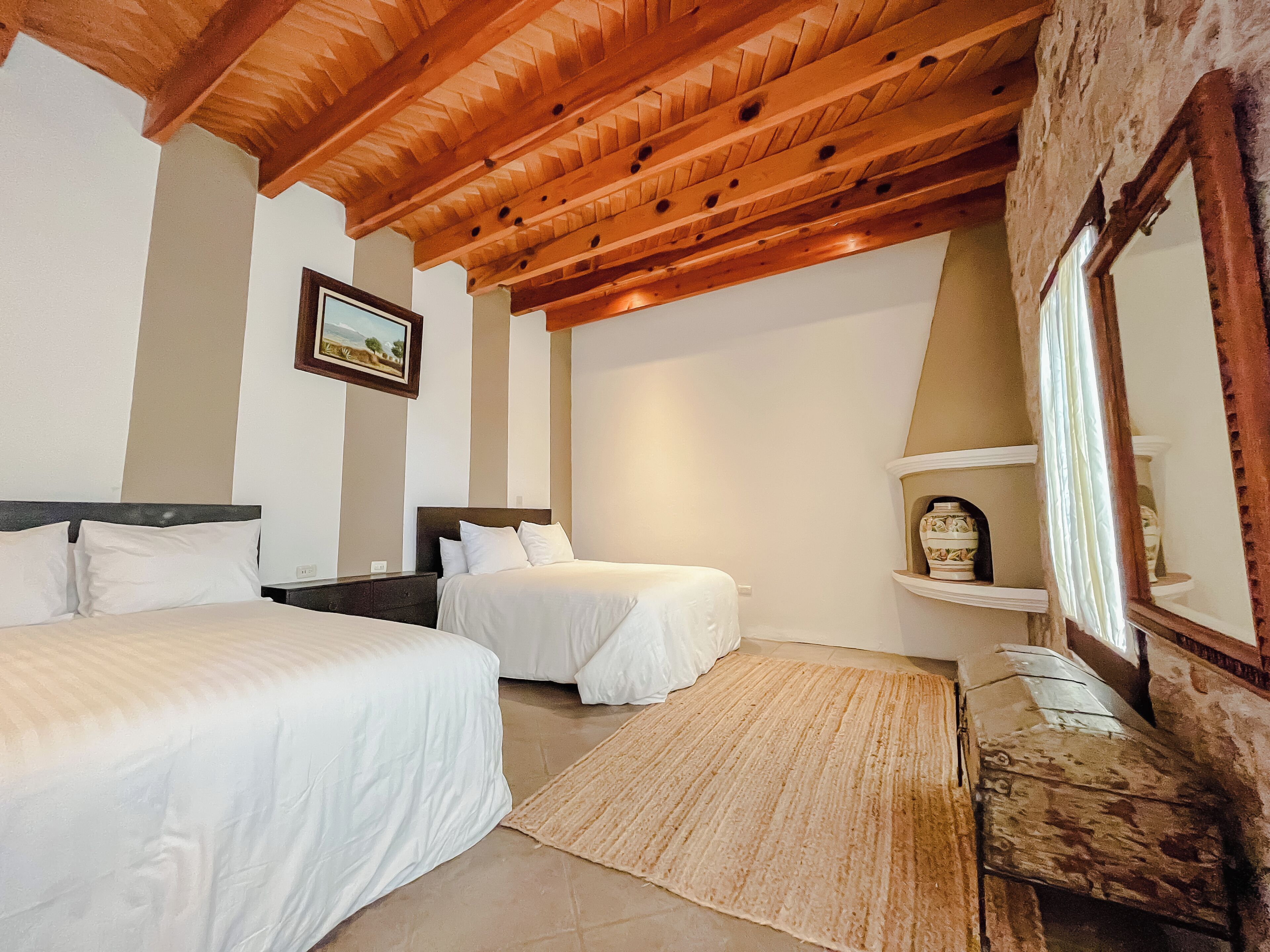 A bedroom with two beds, a wooden ceiling, a painting, a mirror, and a small shelf with decorative items.