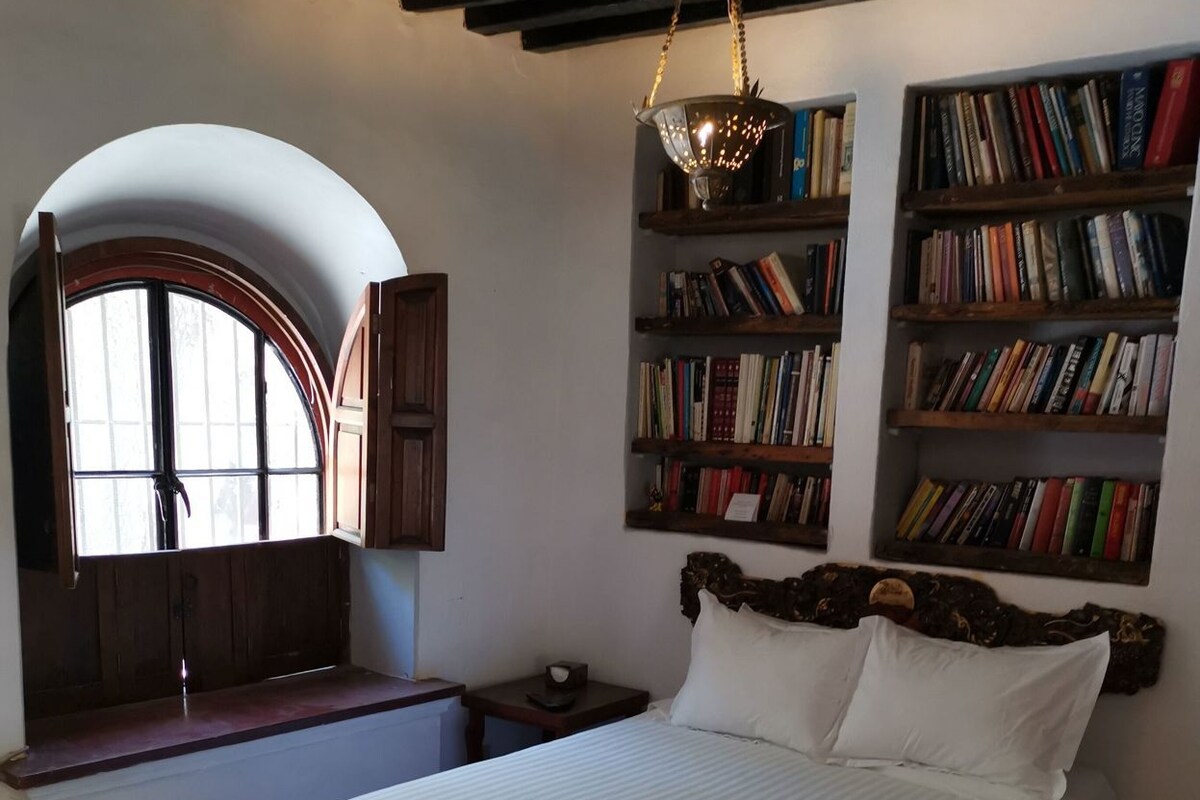 Una camera da letto con un letto grande, una finestra con persiana aperta, una libreria e una lampada a sospensione.