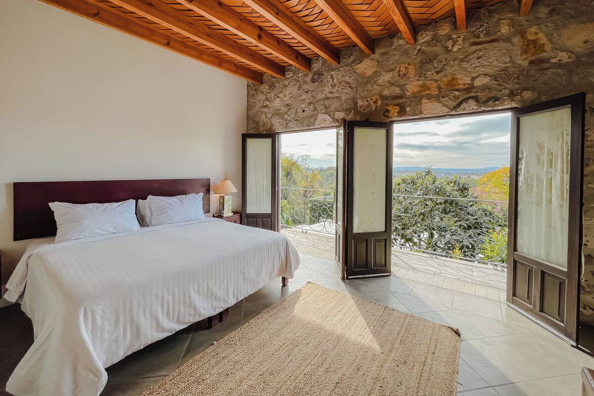 Une chambre avec un mur en pierre, des poutres en bois au plafond, un lit avec une literie blanche et une vue sur un balcon, accessible par une porte ouverte.