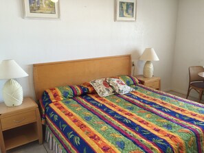 A bedroom with a wooden headboard, a colorful floral bedspread, two bedside lamps, a chair, and two framed pictures on the wall.