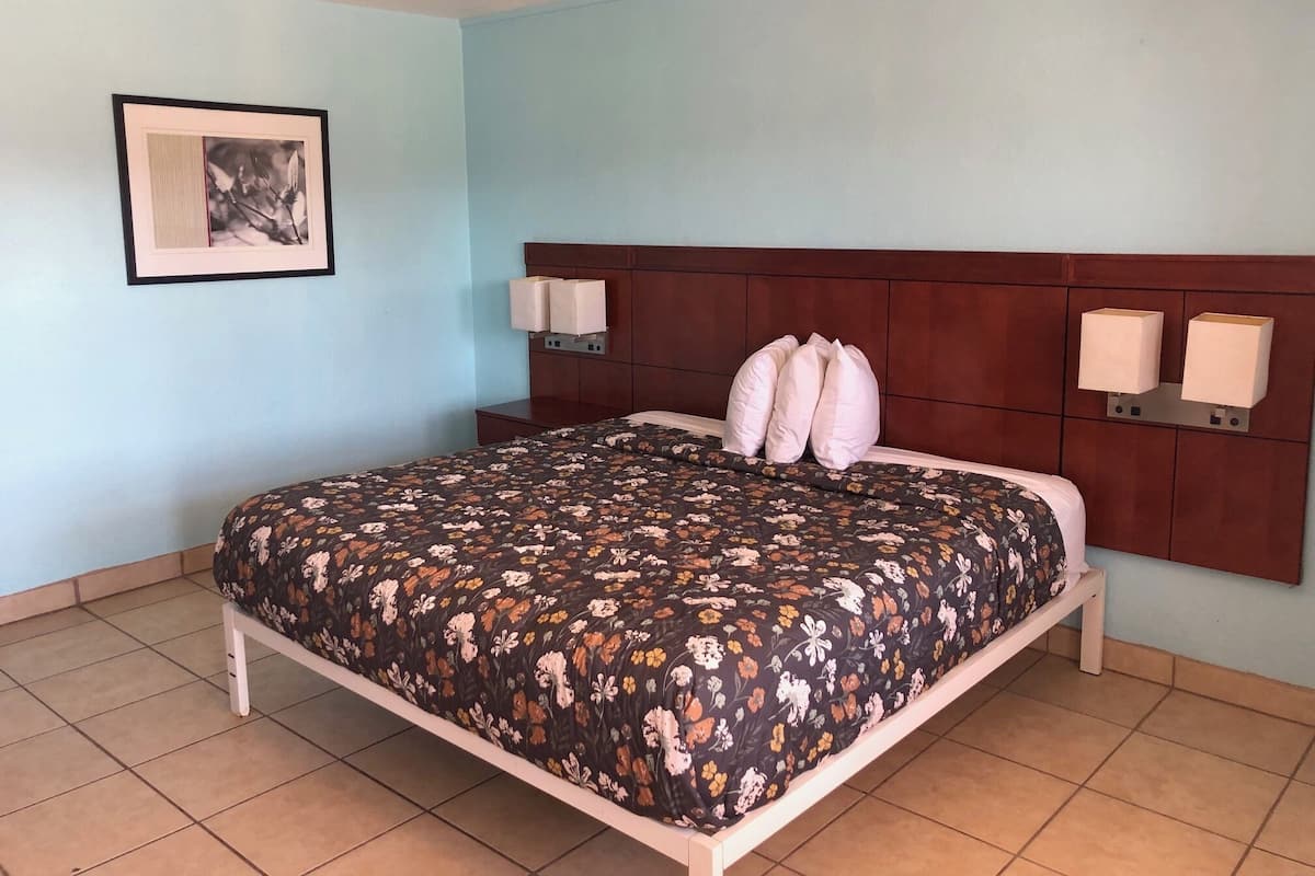 A hotel room with a floral bedspread, a wooden headboard, two wall-mounted lamps, and a framed picture on the wall.