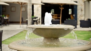 Una fontana in pietra con acqua che zampilla al centro, circondata da posti a sedere all’aperto e ombrelloni.