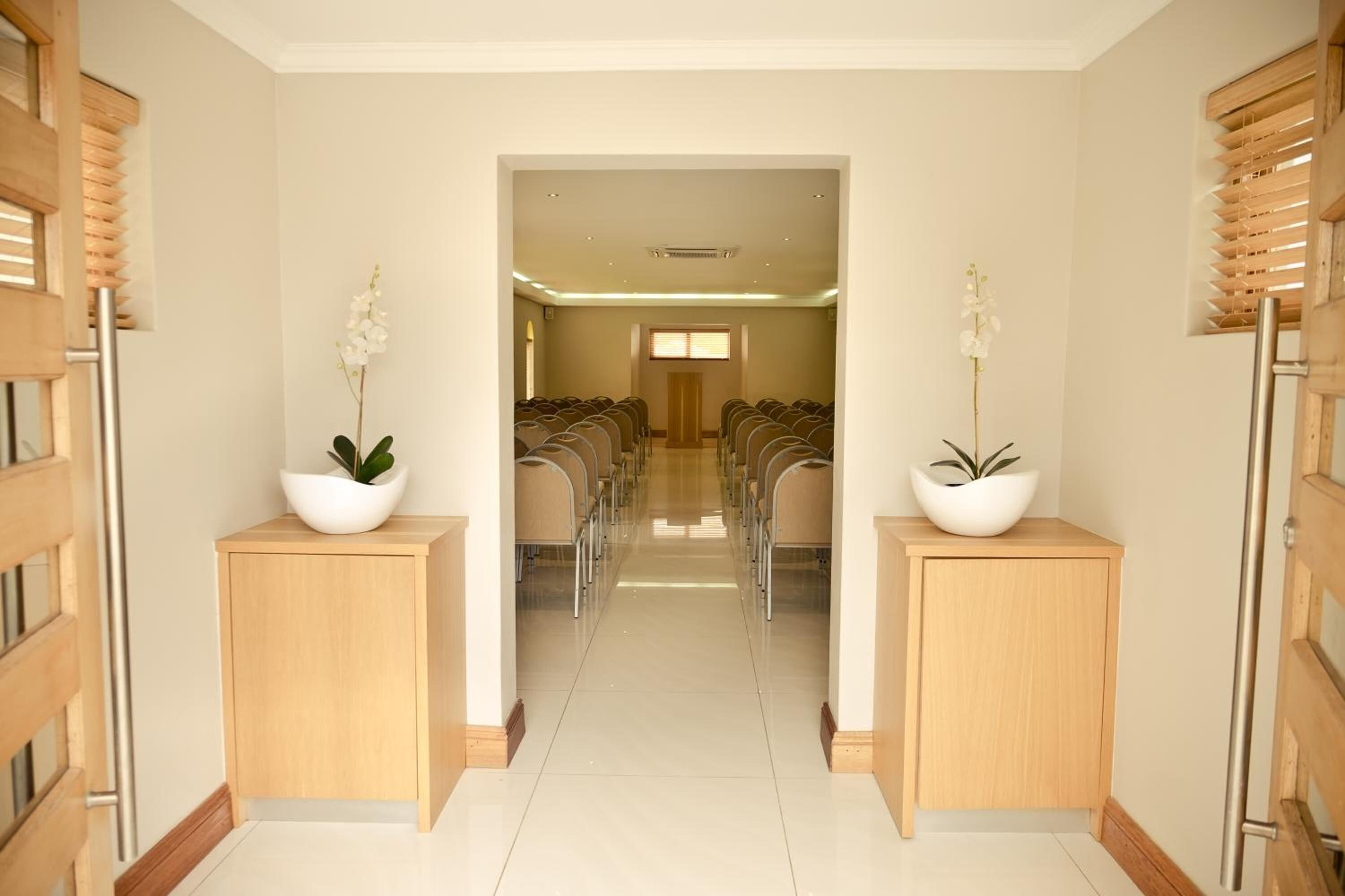 A hotel lobby with a central corridor leading to a seating area, wooden cabinets with potted plants, and a tiled floor.