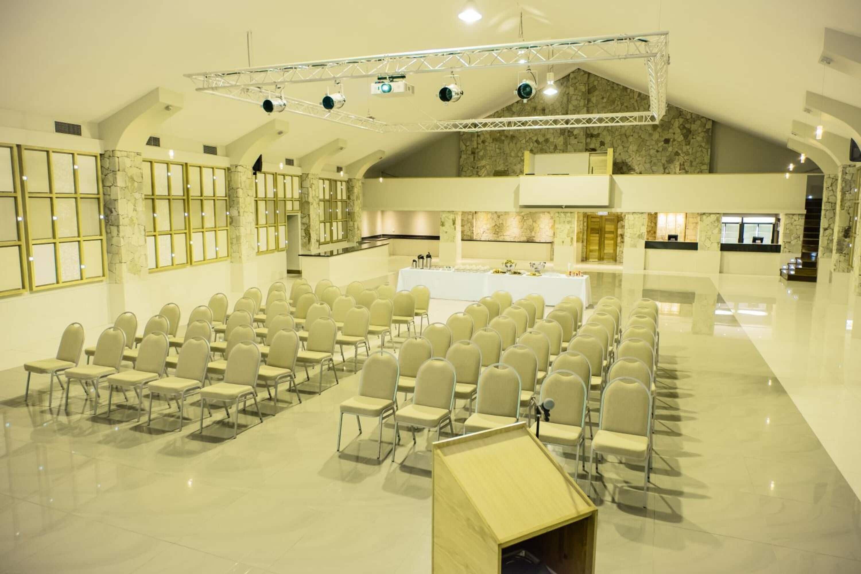 Un salón de conferencias amplio con filas de sillas, un escenario con iluminación y una zona de cocina al fondo.