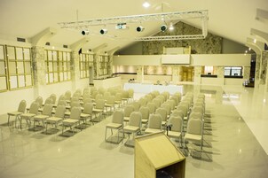 Un salón de conferencias amplio con filas de sillas, un escenario con iluminación y una zona de cocina al fondo.