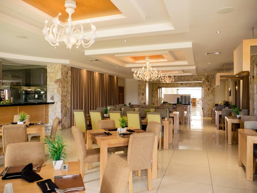 Een ruime hotel eetzaal met meerdere tafels en stoelen, een bar in het midden en uitzicht op de keuken.
