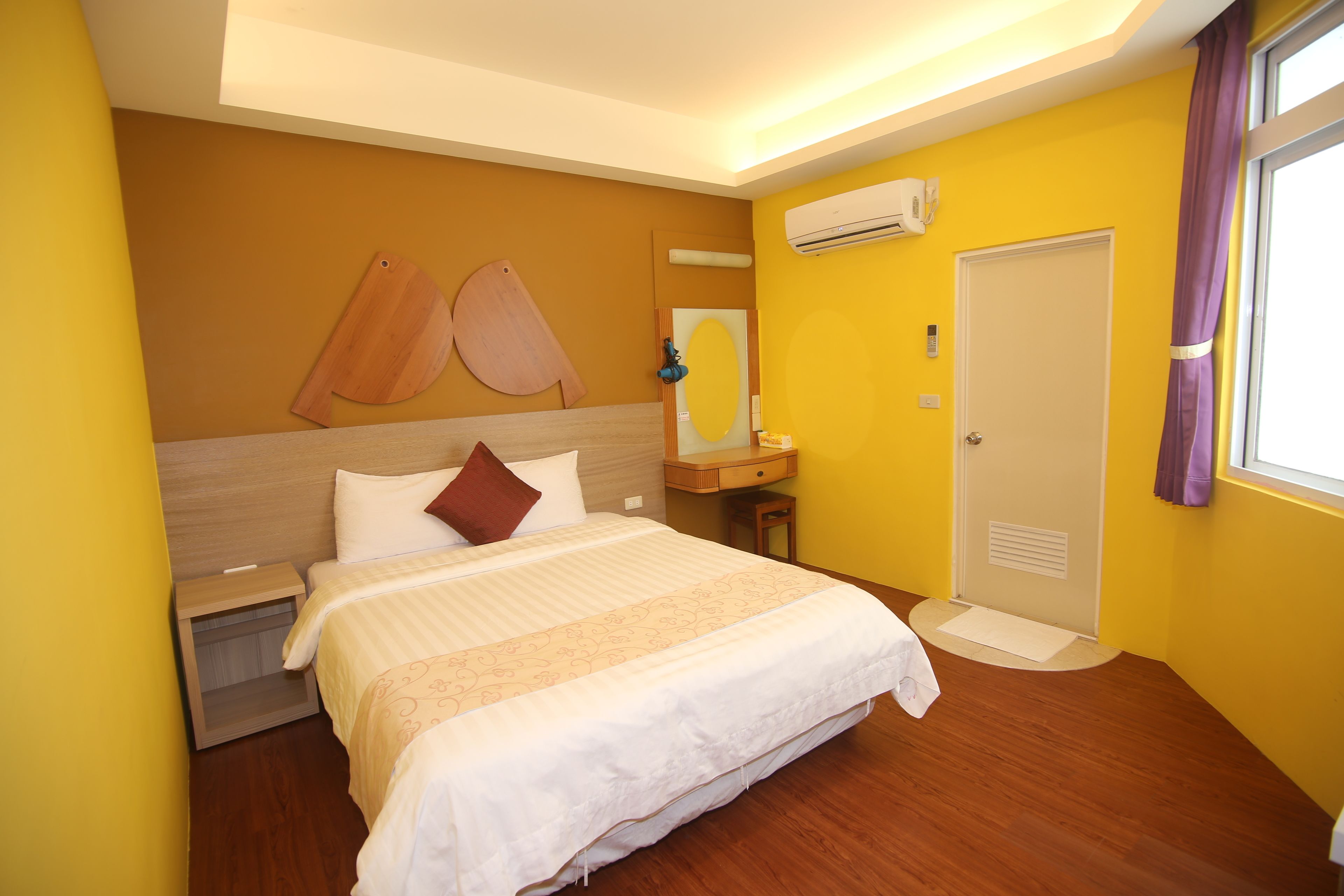 A hotel room with a bed, a nightstand, a mirror, an air conditioning unit, a door, and a window with curtains.