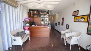 Uma área de recepção com um balcão de madeira, cadeiras de vime branco e uma parede decorativa com obras de arte emolduradas.