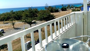 Varanda com mesa e cadeiras de vidro, com vista para uma praia, palmeiras e céu azul límpido.