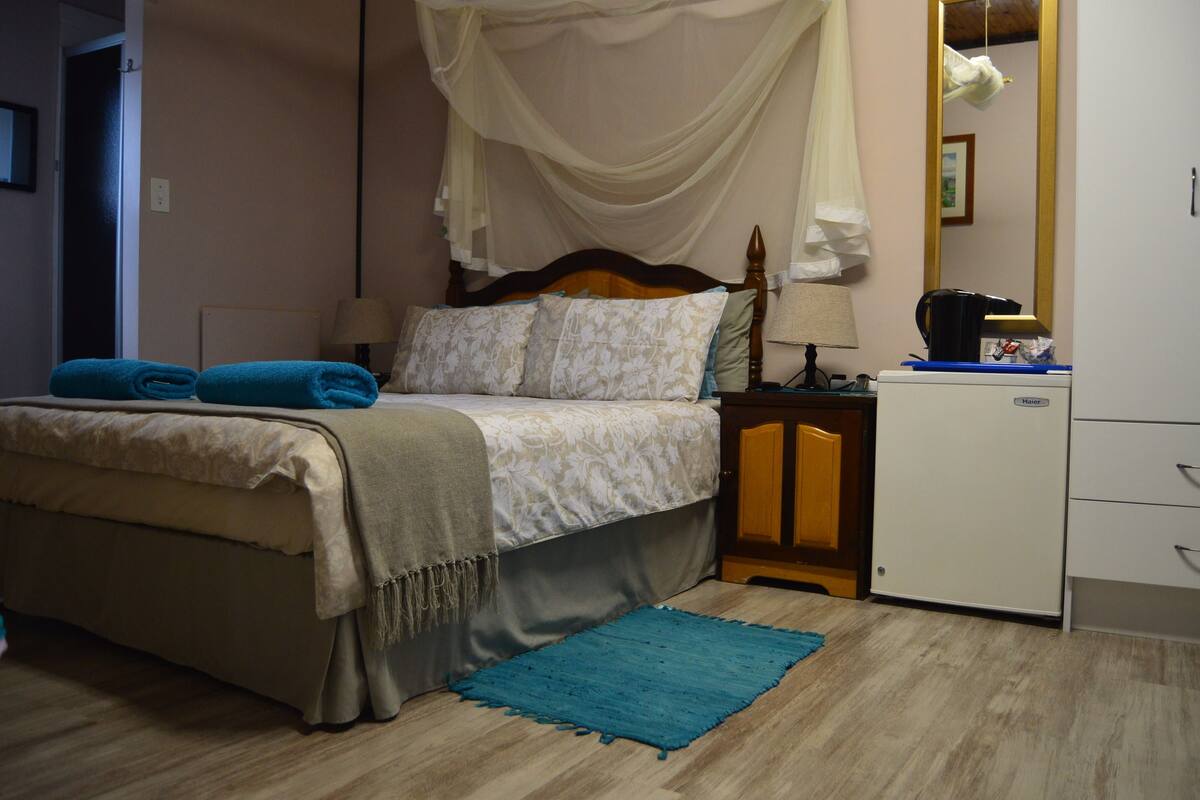 A bedroom with a bed, a nightstand, a desk, a mirror, and a small refrigerator.